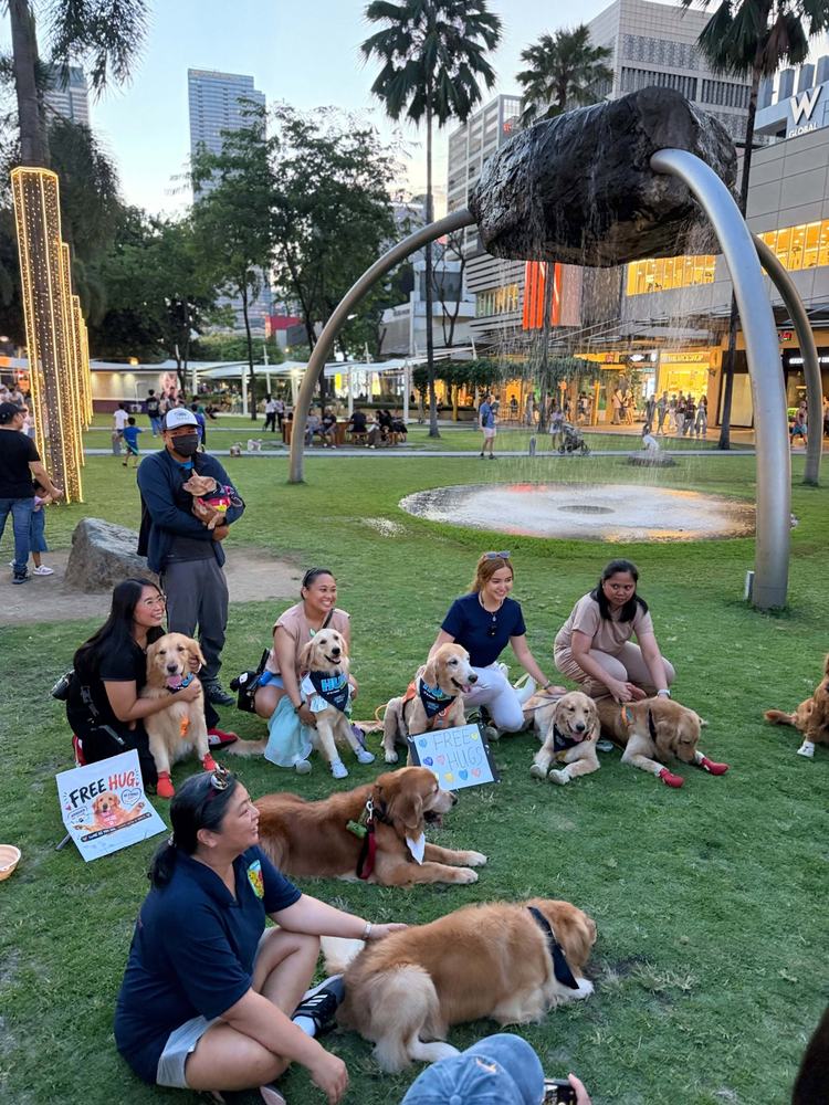 Members and their golden retrievers sitting in a circle on the lawn at BGC with Free Hugs signs